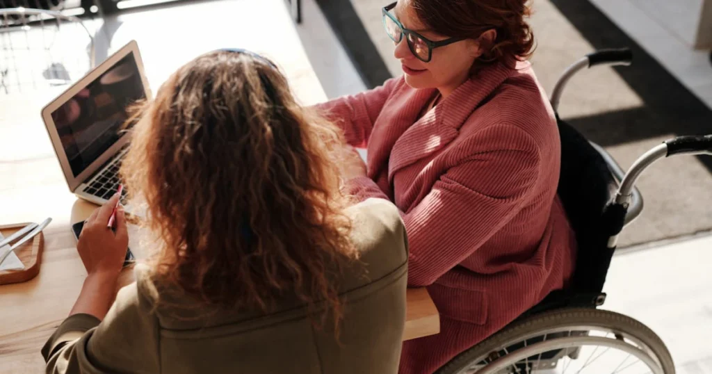 Duas mulheres sentadas em uma mesa de trabalho olhando um notebook uma delas está em uma cadeira de rodas representando o auxílio-acidente, que permite que o beneficiário continue trabalhando